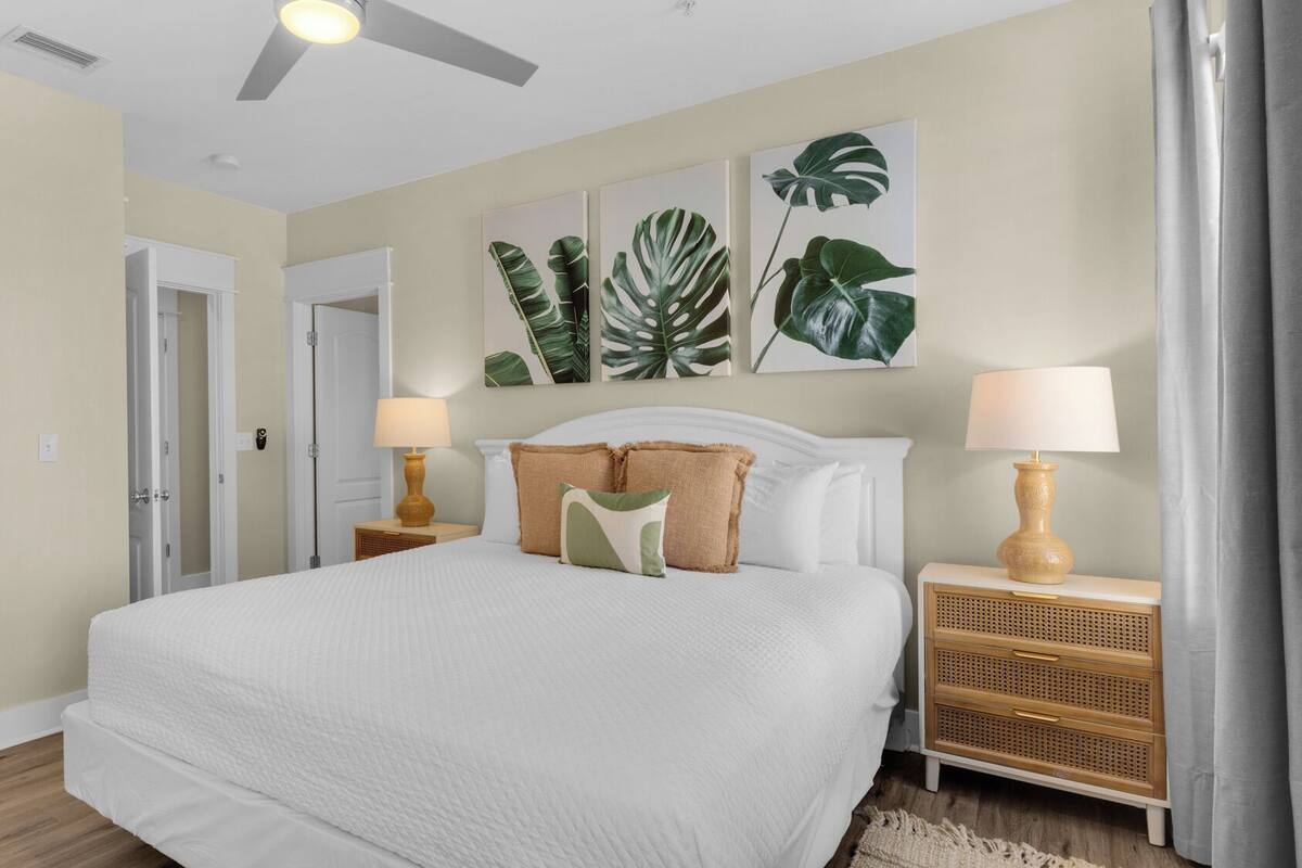 A bedroom with a large bed, two bedside tables with lamps, a ceiling fan, and tropical leaf wall art.