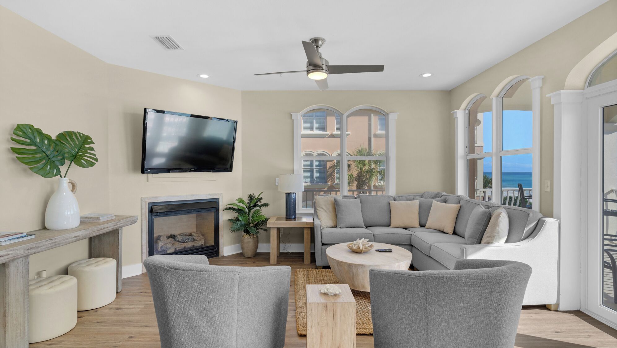 A spacious living room with a grey sectional sofa, a flat-screen TV mounted on the wall, a fireplace, and large windows offering a view of the ocean.