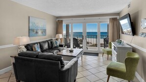A living room with a black leather sofa, a green armchair, and a dining table. There is a painting on the wall and a TV mounted on the wall. The room has a view of the ocean through large glass doors. - Moondrifter Condominiums (Panama City Beach)