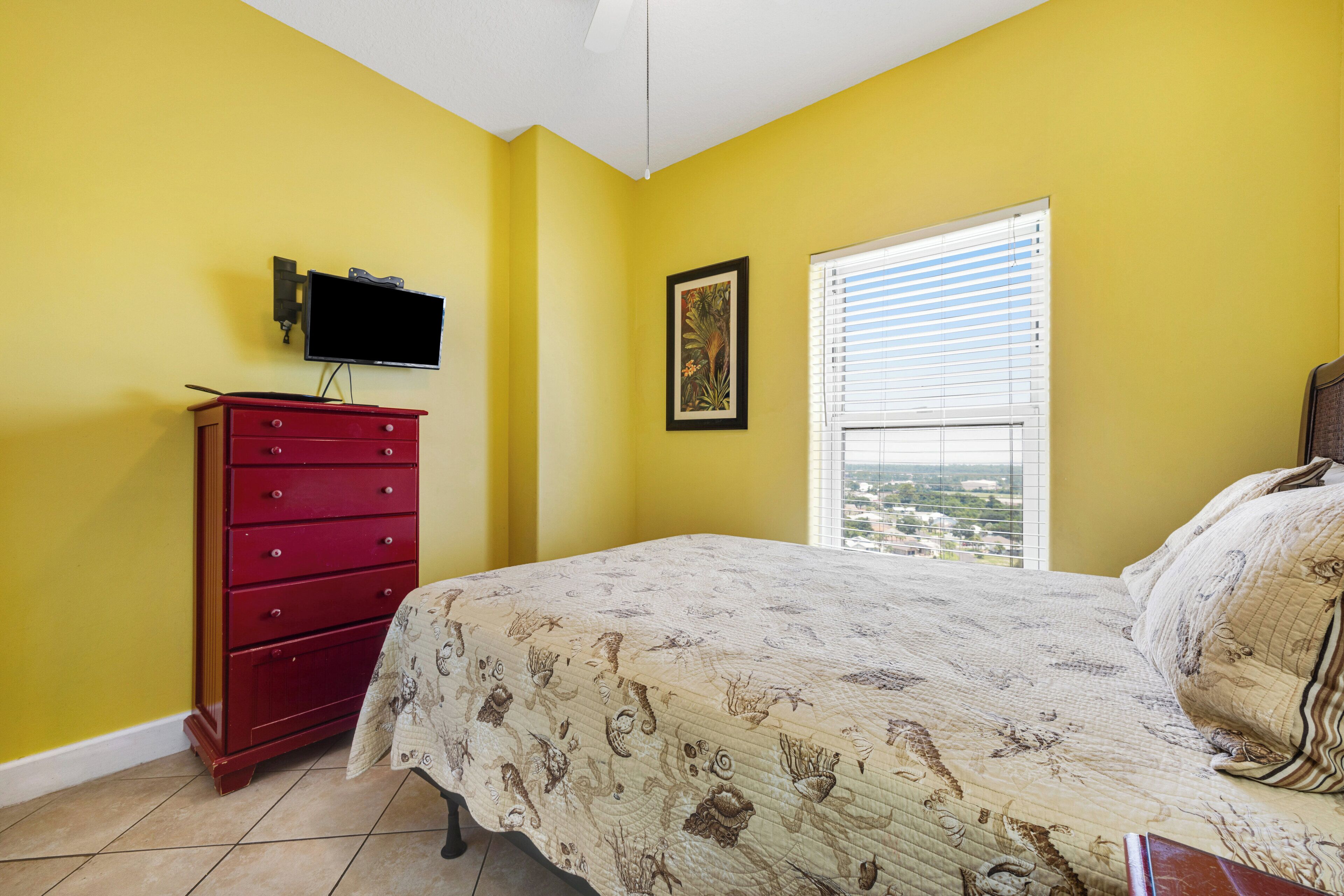 A bedroom with a bed, a red dresser, a wall-mounted TV, and a window with blinds.