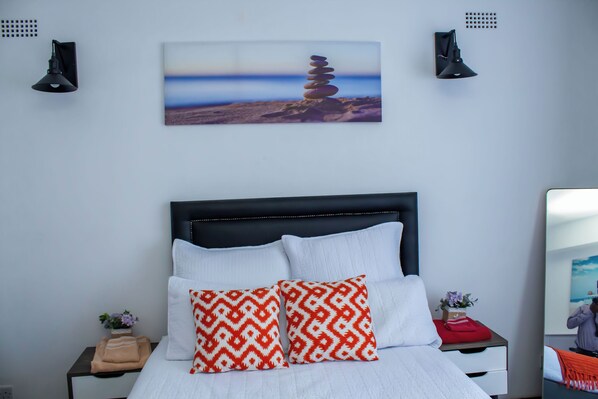 Una cama bien hecha con almohadillas blancas y anaranjadas, un cabecero con detalles decorativos y un cuadro en la pared con una pila de piedras.