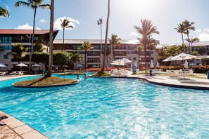 A swimming pool with palm trees and umbrellas, surrounded by a hotel building.