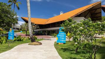 A modern building with a sloped roof, surrounded by greenery and palm trees.