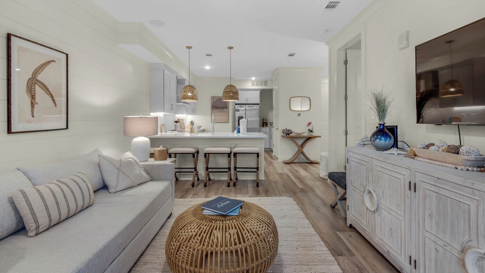 A modern living room with a sofa, a round wicker ottoman, and a dining area with a kitchen in the background.