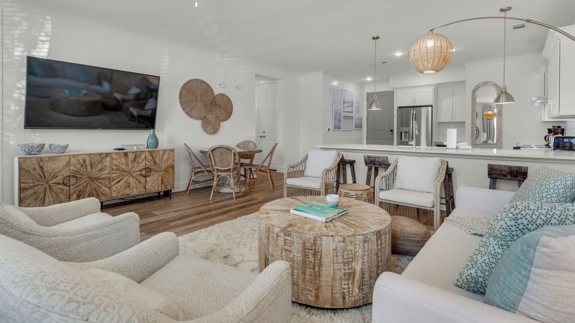 A modern living room with a large flat-screen TV, wicker furniture, a round coffee table, and a kitchen area with white cabinets and a large mirror.
