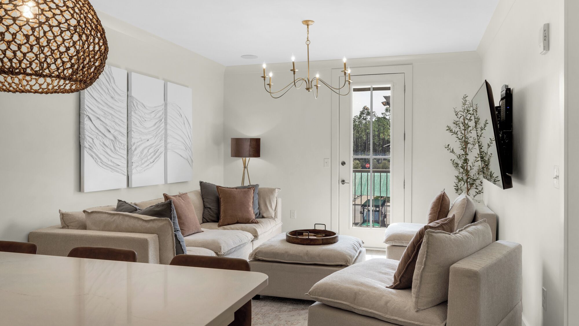 A modern living room with a dining area, featuring a chandelier, artwork, and a flat-screen TV mounted on the wall.