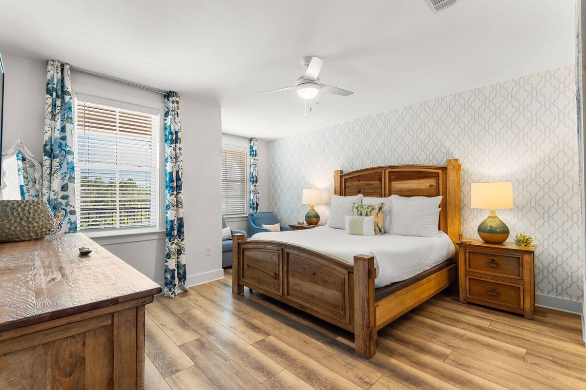 A bedroom with a wooden bed, two bedside tables with lamps, a ceiling fan, and a window with blinds.