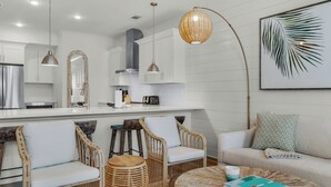 A modern kitchen with white cabinetry, a central island, and pendant lighting. There are wicker chairs and a sofa in the living area.