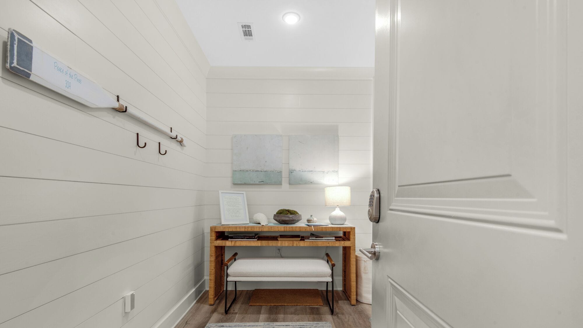 A hallway with white paneling, a wooden bench, a small table with a lamp, and a door with a keypad lock.