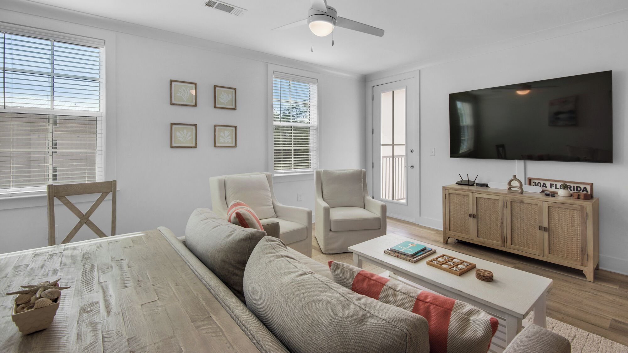 A modern living room with a sofa, armchairs, a coffee table, and a TV. There are framed pictures on the wall, a wooden cabinet, and a window with blinds.