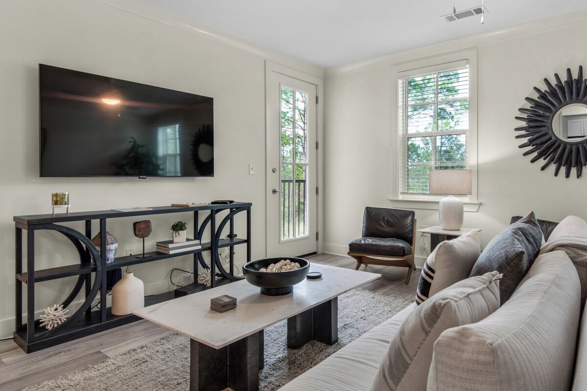 A living room with a large flat-screen TV, a dark console table with decorative items, a white coffee table, a leather armchair, and a gray sofa.