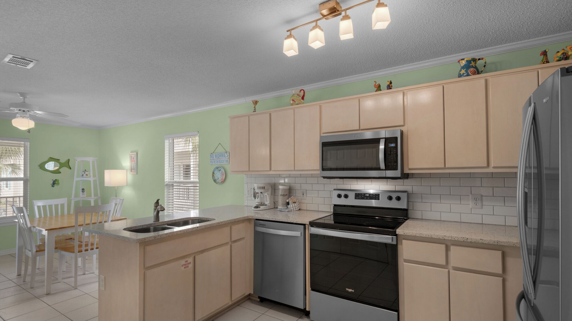 A modern kitchen with light green walls, beige cabinets, a stainless steel refrigerator, a microwave, an oven, and a dishwasher. There is a dining area with white chairs and a wooden table.