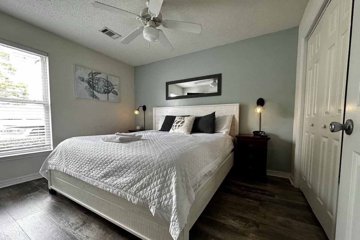 A bedroom with a bed, a ceiling fan, a nightstand, a bedside lamp, a door, and a window with blinds.