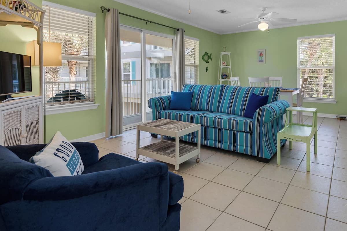 A living room with a striped sofa, a coffee table, and a TV. There is a balcony with a view, and a kitchen area with a bar stool.