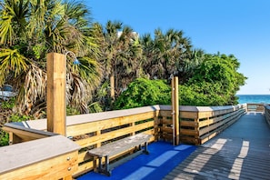 A wooden boardwalk with benches, surrounded by lush greenery and palm trees, leading to a beach. - Rollin Tide Villas (Santa Rosa Beach)