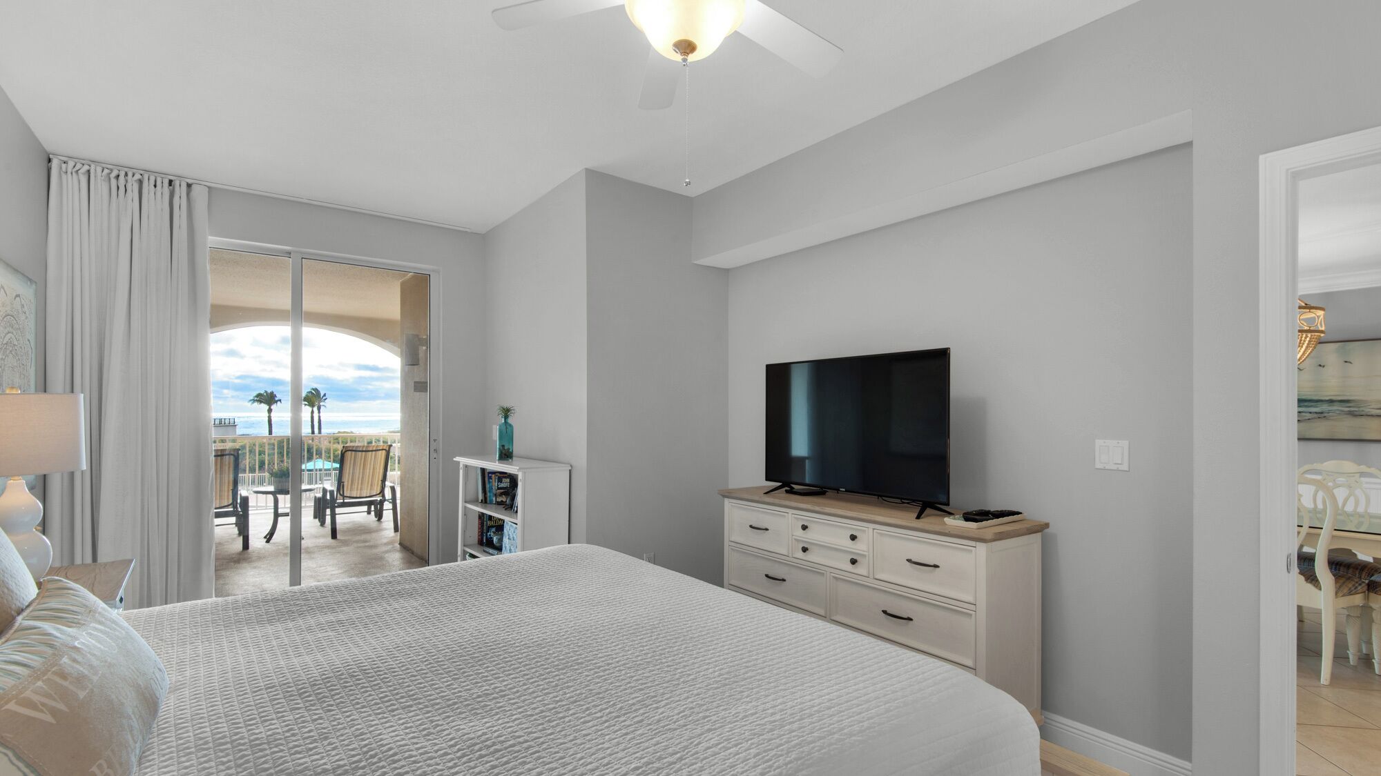 A bedroom with a large bed, a TV on a dresser, a balcony with chairs, and a view of the ocean.