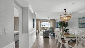 A spacious dining area with a glass table, white chairs, and a chandelier. - San Remo (Santa Rosa Beach)