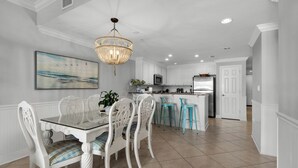 A modern kitchen with white cabinetry, a central island, and a dining area with a glass table and white chairs. - San Remo (Santa Rosa Beach)
