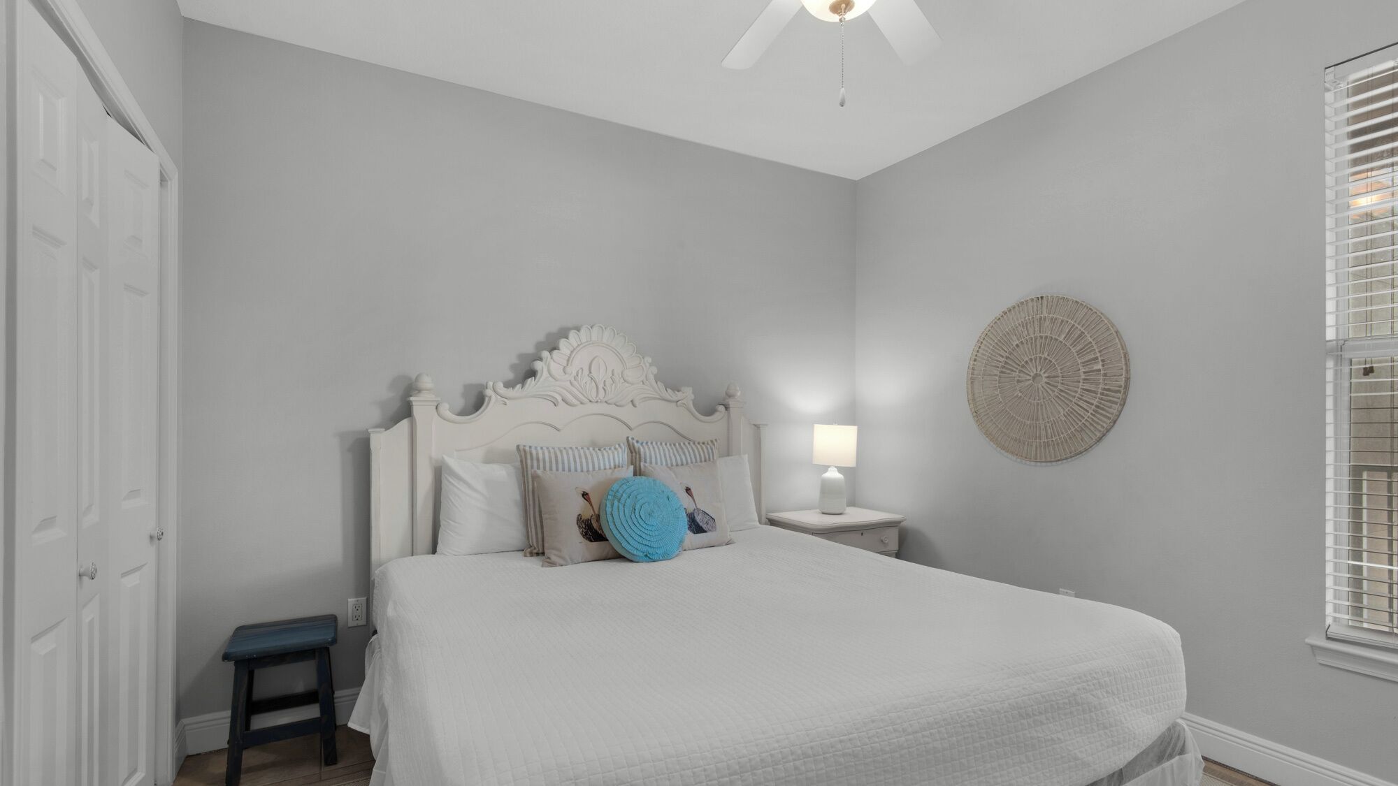 A bedroom with a white bed, a nightstand with a lamp, a small table, and a decorative wall piece.