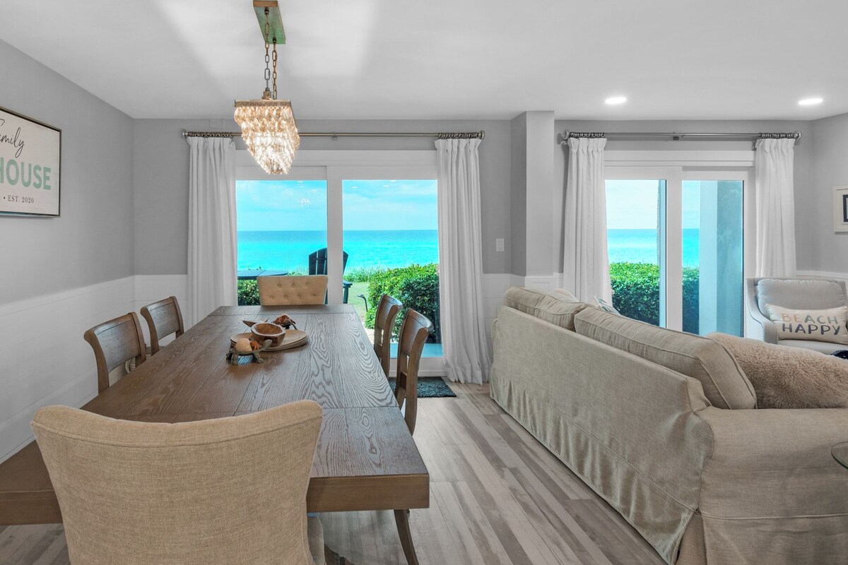 A dining area with a view of the ocean, a beach-themed sign, and a cozy seating area.
