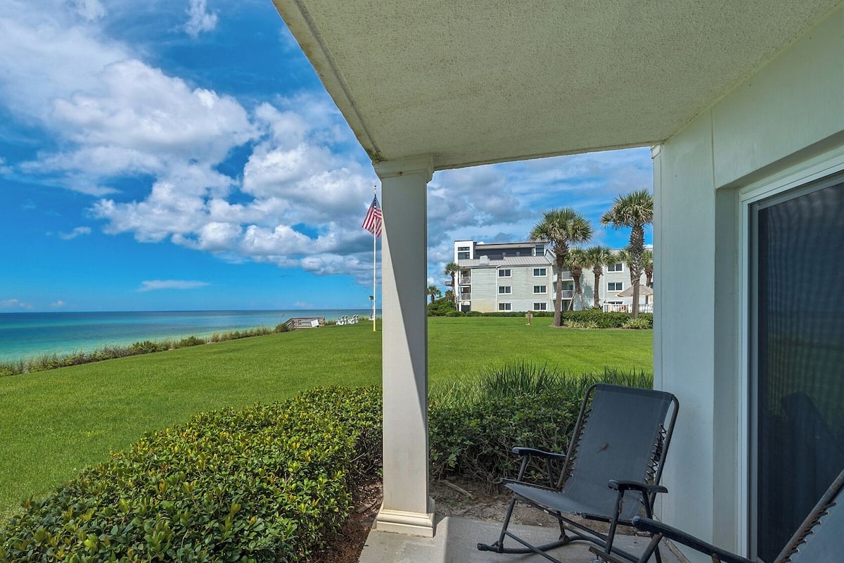 A covered outdoor area with a view of the ocean, a lawn, and a building with multiple windows and balconies.