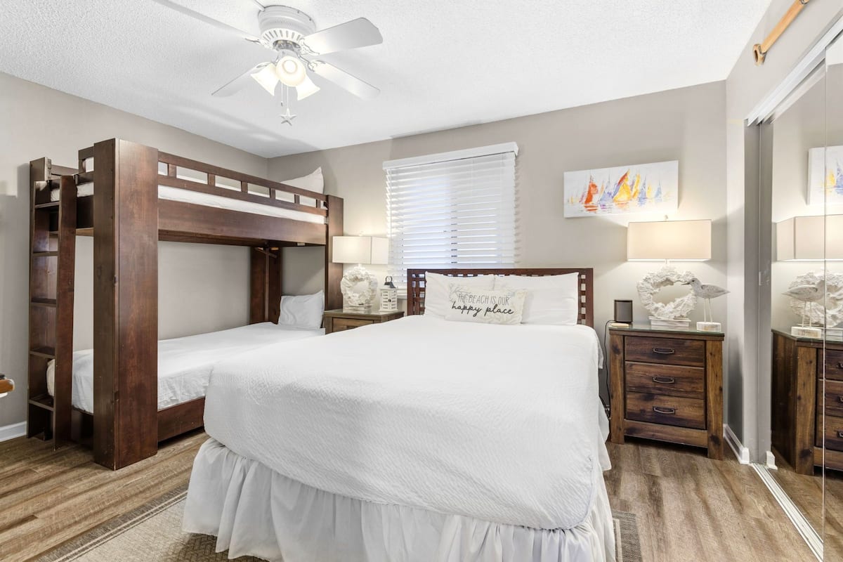 A bedroom with a bunk bed, a single bed, a ceiling fan, a bedside table with a lamp, a nightstand, and a wall with a framed picture.