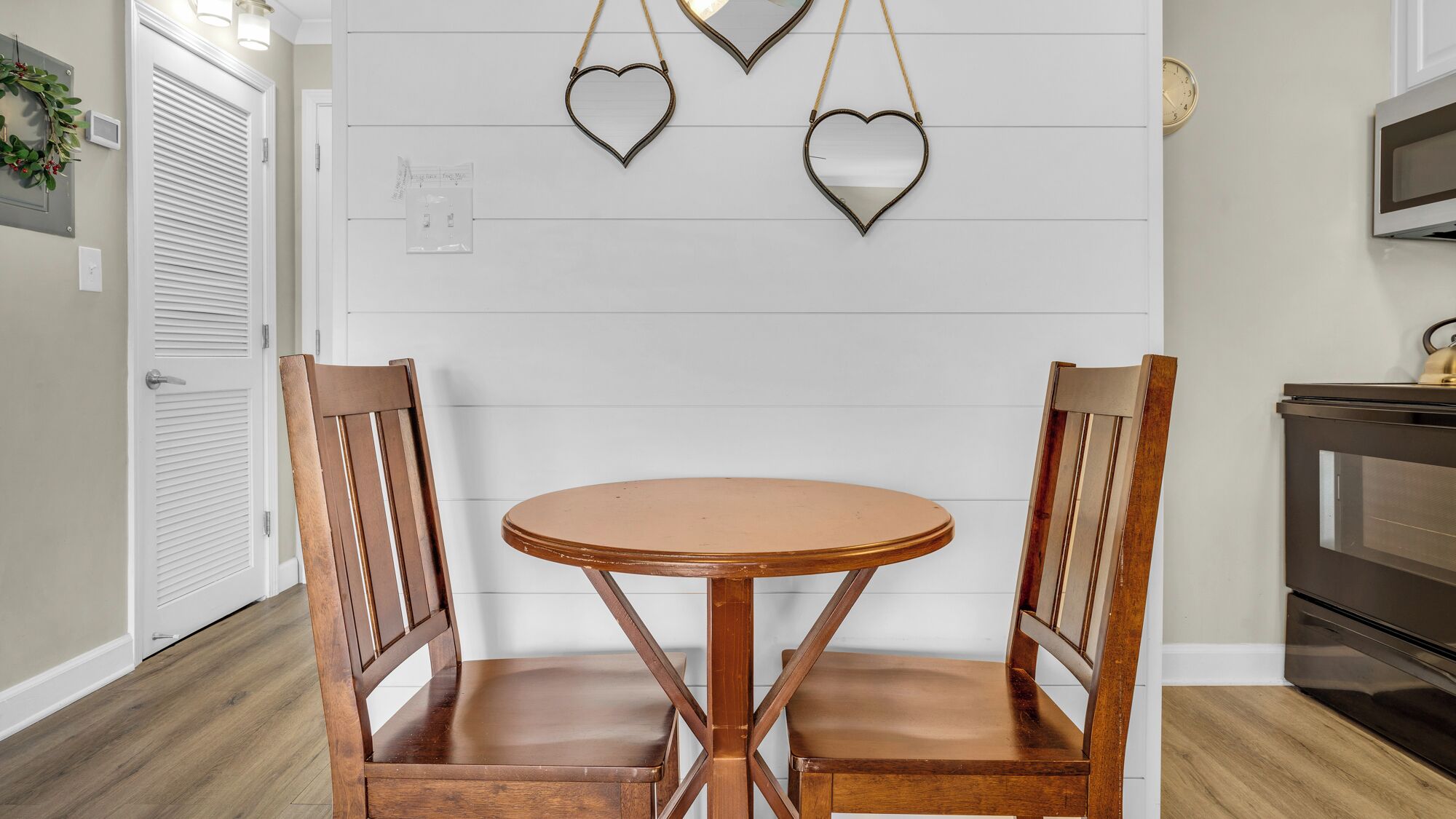 Un coin repas avec une table ronde et deux chaises en bois, un mur blanc orné de panneaux horizontaux et un miroir décoratif en forme de cœur accroché au-dessus.