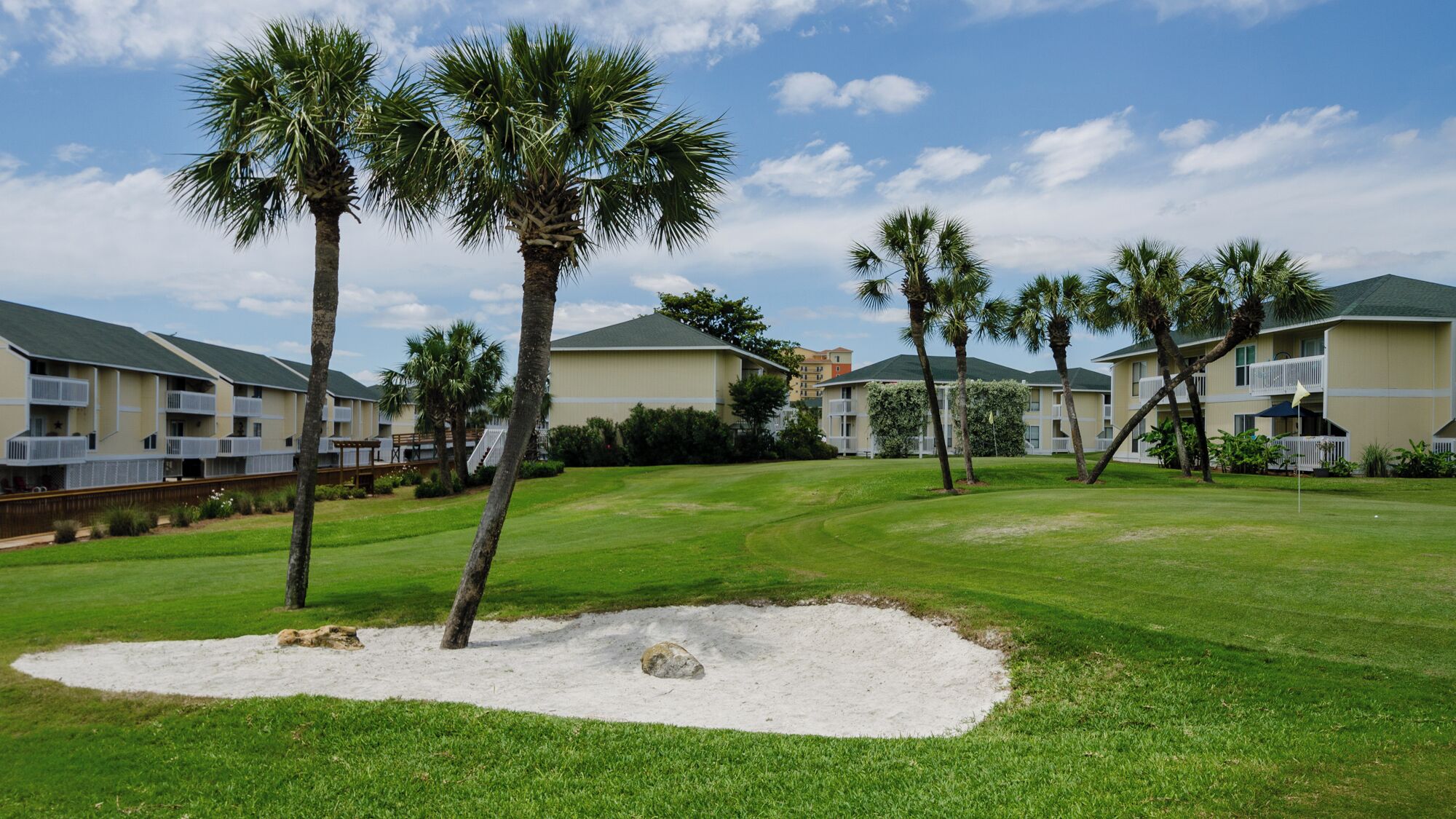 Un campo de golf con bunkers de arena y palmeras, rodeado de edificios de varios pisos con balcones.