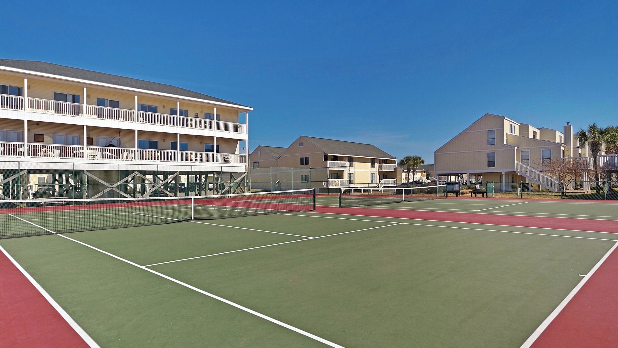 A tennis court with a net in the middle, surrounded by buildings with balconies and a clear blue sky.