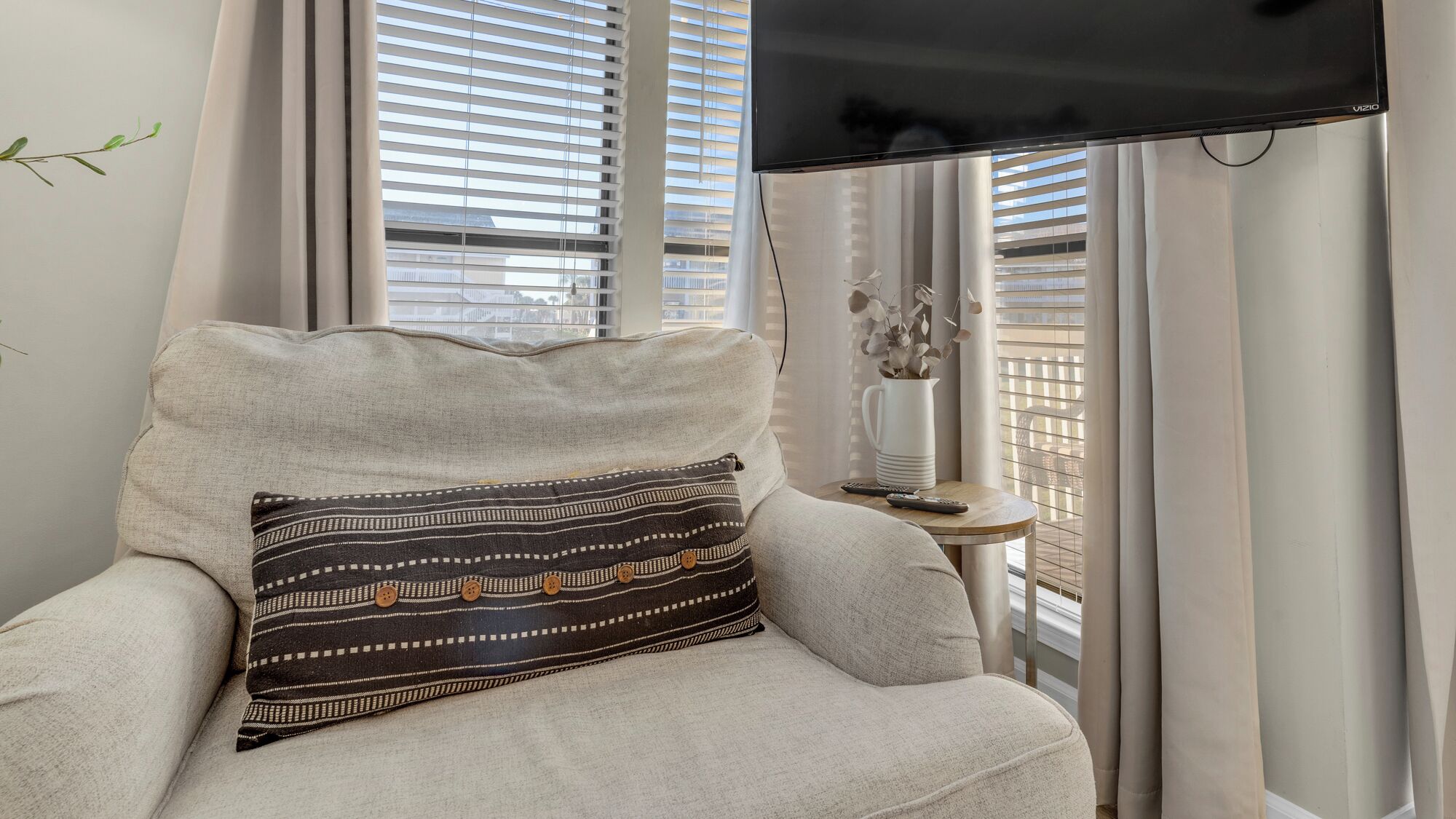 A modern living room with a beige armchair, a patterned throw pillow, and a flat-screen TV mounted on the wall.