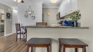 Cocina con encimera de granito, armarios blancos y una lámpara colgante. Hay dos taburetes de madera y una cesta tejida sobre la encimera.