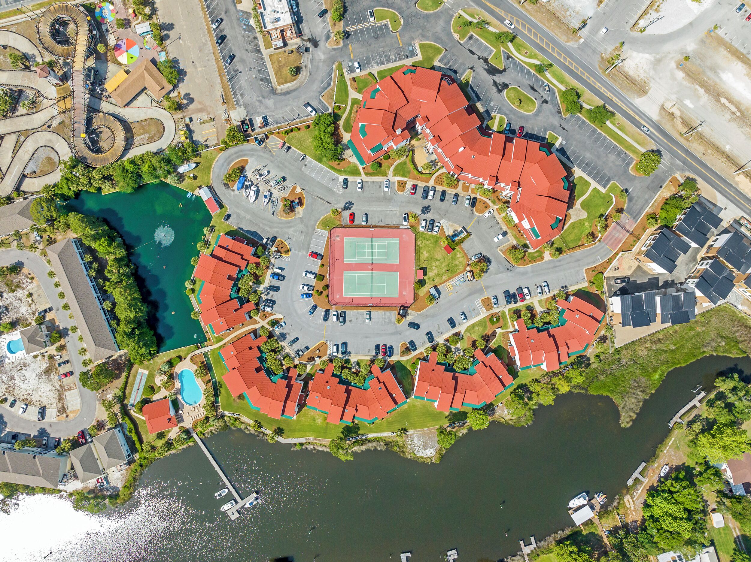 An aerial view of a residential area with a central sports court, surrounding houses, and a body of water.