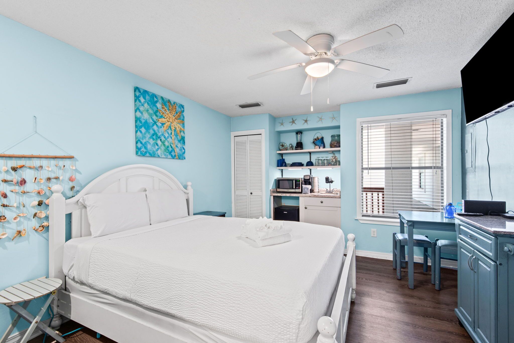 A bedroom with a bed, a ceiling fan, a desk with a chair, and a window with blinds.