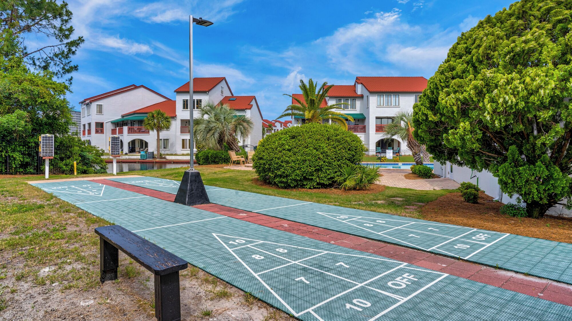 Un terrain de jeu de shuffleboard avec un banc, un lampadaire et une piscine en arrière-plan.
