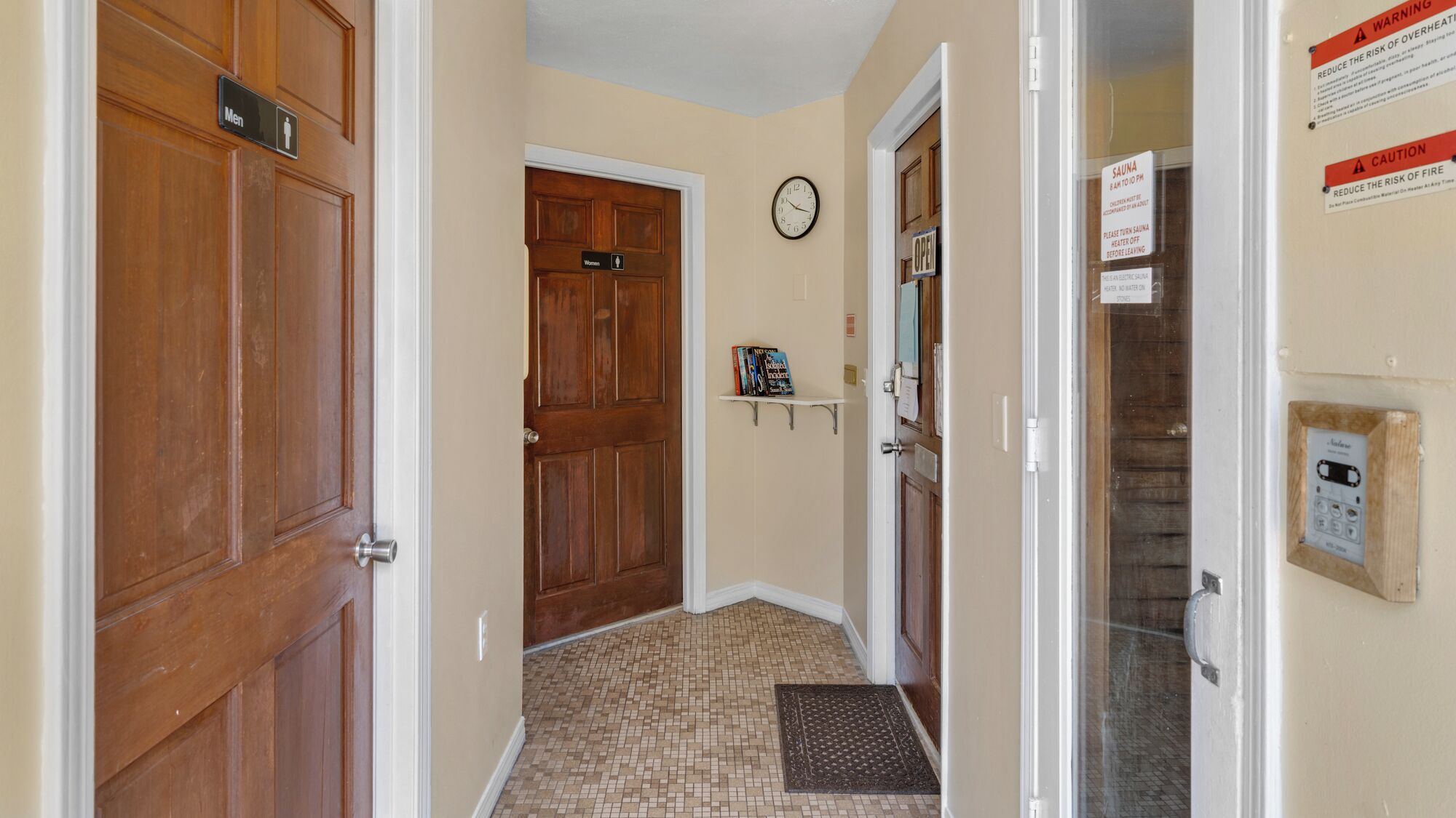 A hallway with wooden doors labeled "Men" and "Women," a clock on the wall, and a sign indicating the sauna is open.