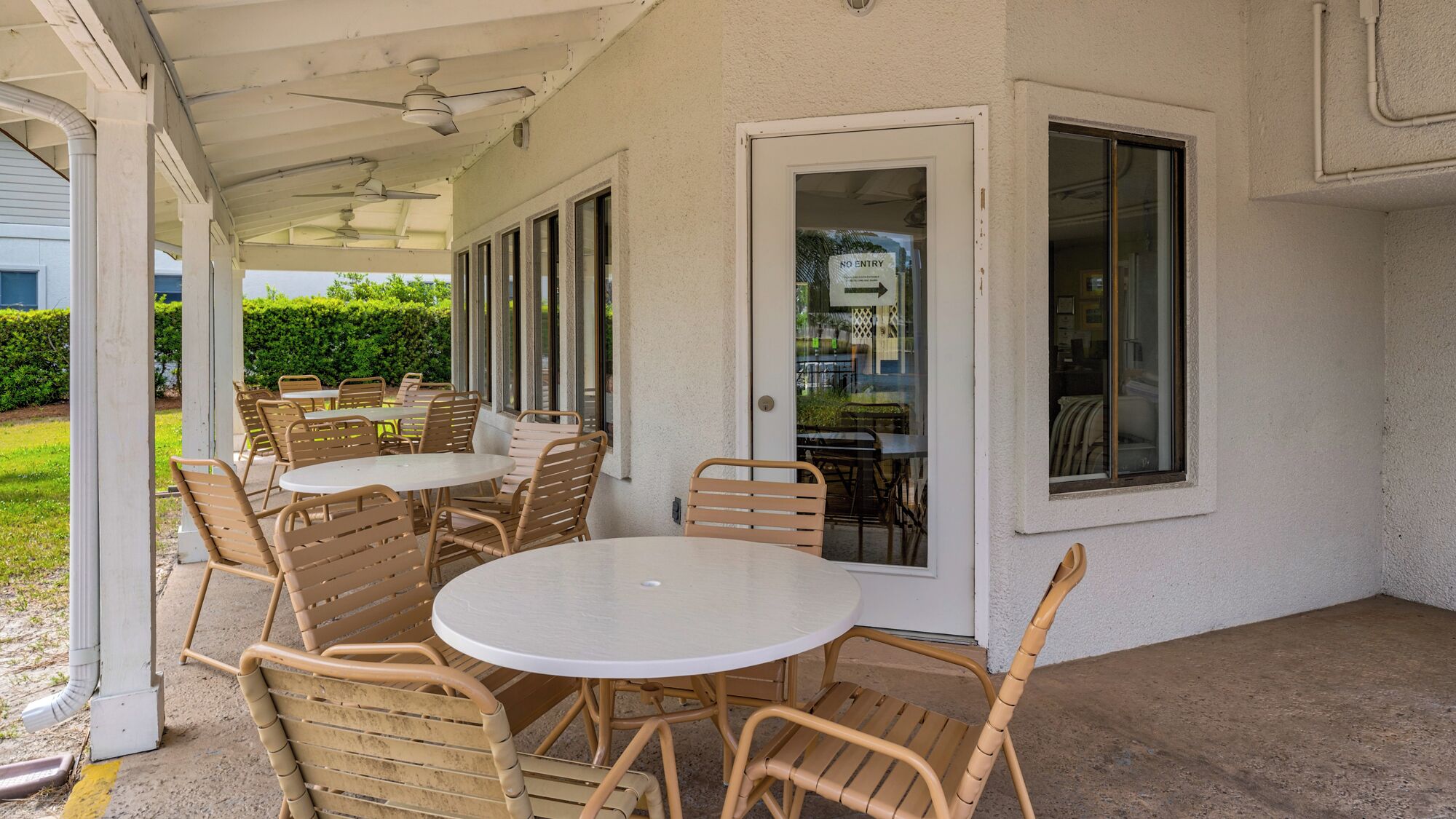 Une terrasse extérieure avec des tables rondes et des chaises en osier, un espace couvert doté de ventilateurs de plafond et une porte vitrée donnant sur l’intérieur.