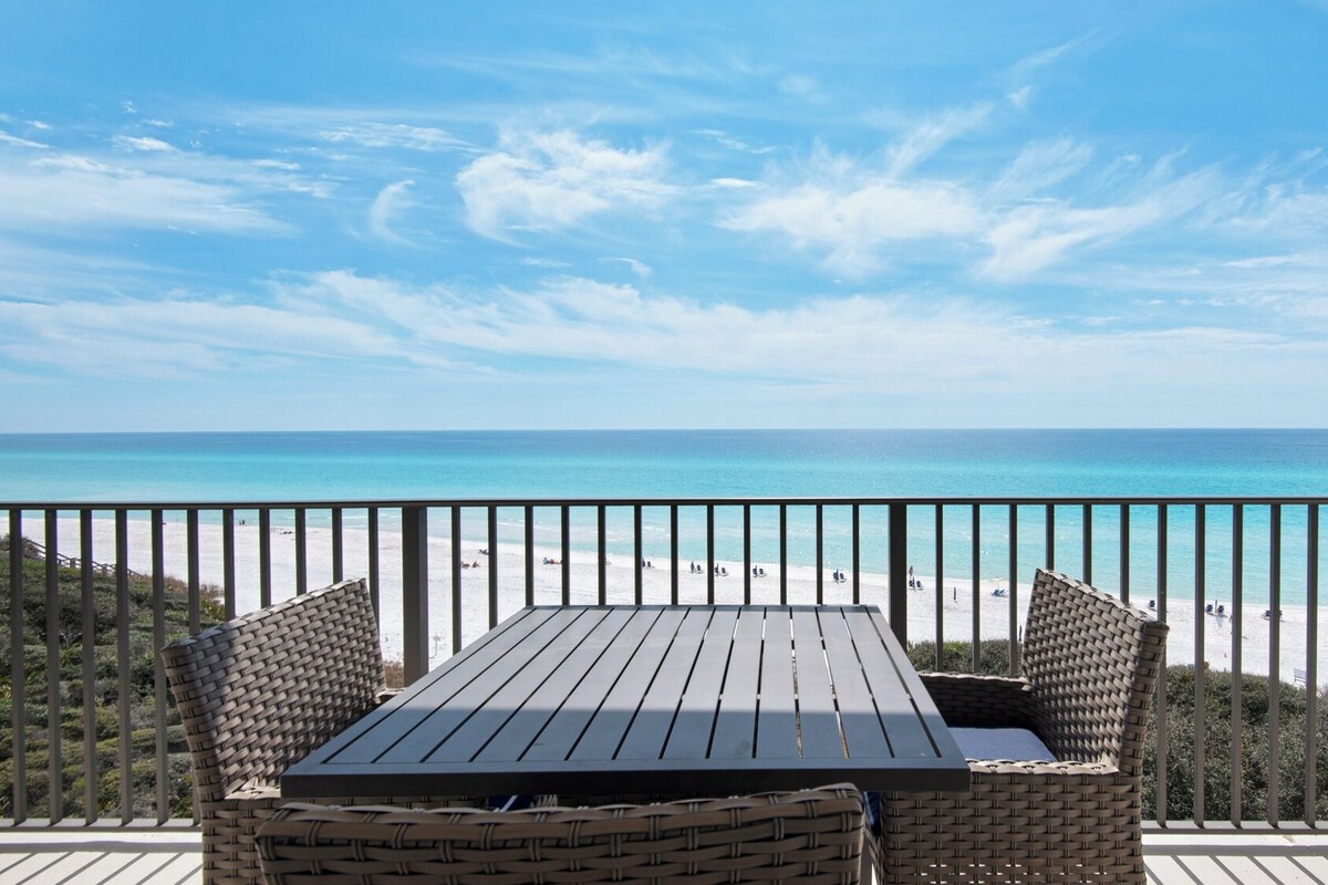 A wooden table and wicker chairs on a balcony overlooking a beach with clear blue water and a sandy shore.
