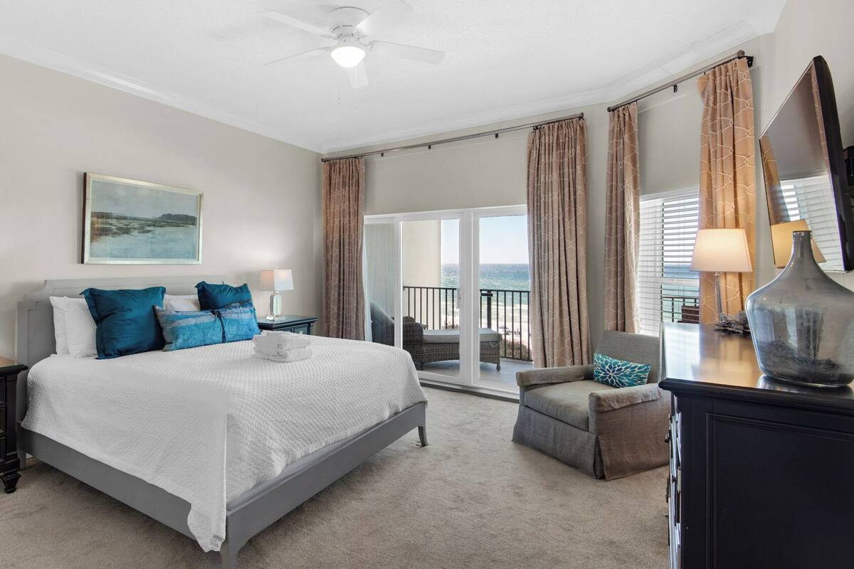 A bedroom with a large bed, a nightstand, a dresser, a chair, and a balcony with curtains.