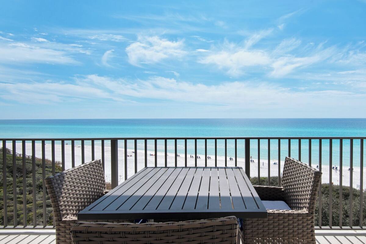 A wooden table and wicker chairs on a balcony overlooking a beach with clear blue water and a sandy shore.