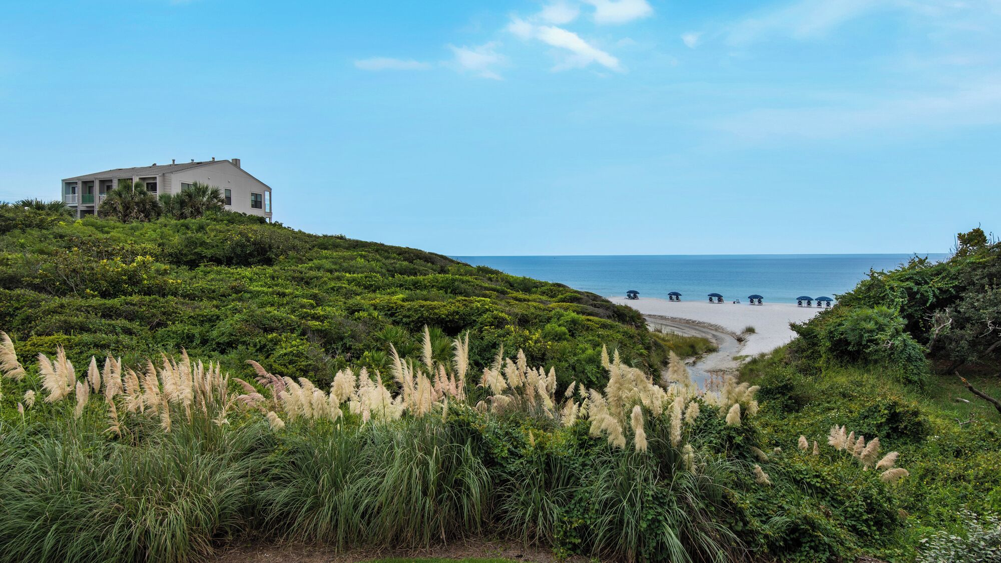 A coastal landscape with a house on a hill overlooking a beach with umbrellas.