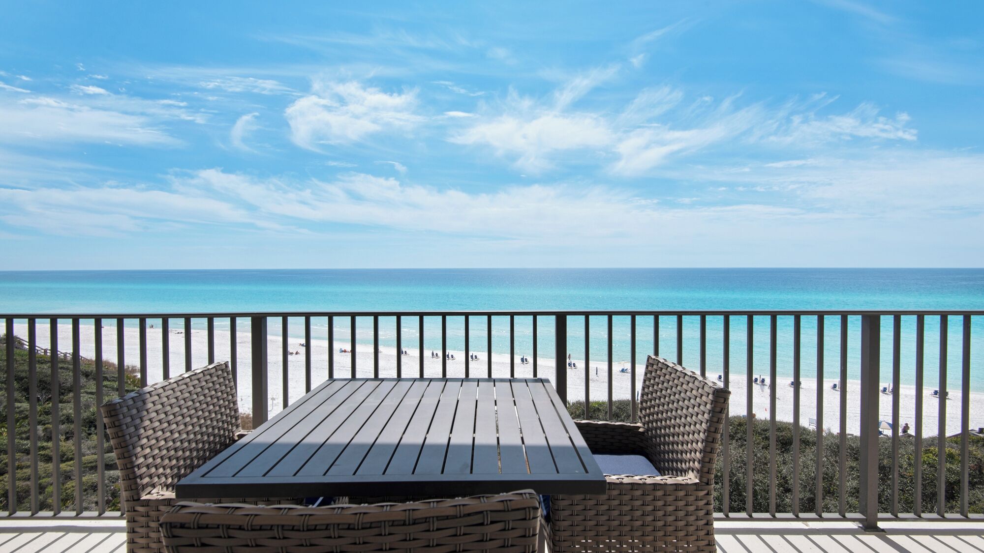 Una mesa de madera y sillas de mimbre en un balcón con vistas a una playa de aguas azules y arena.