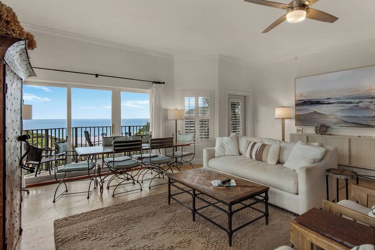 A spacious living room with a large window overlooking the ocean, a ceiling fan, a white sofa, and a dining area with metal chairs.