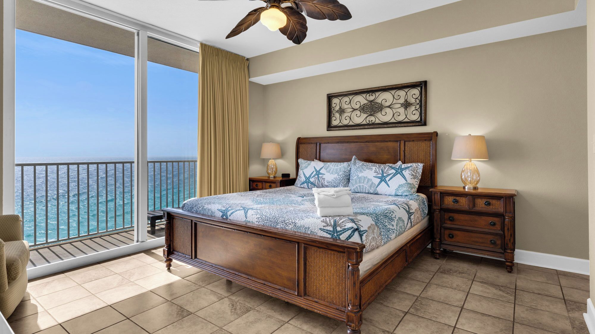 A bedroom with a large bed, two bedside tables, a ceiling fan, and a view of the ocean.