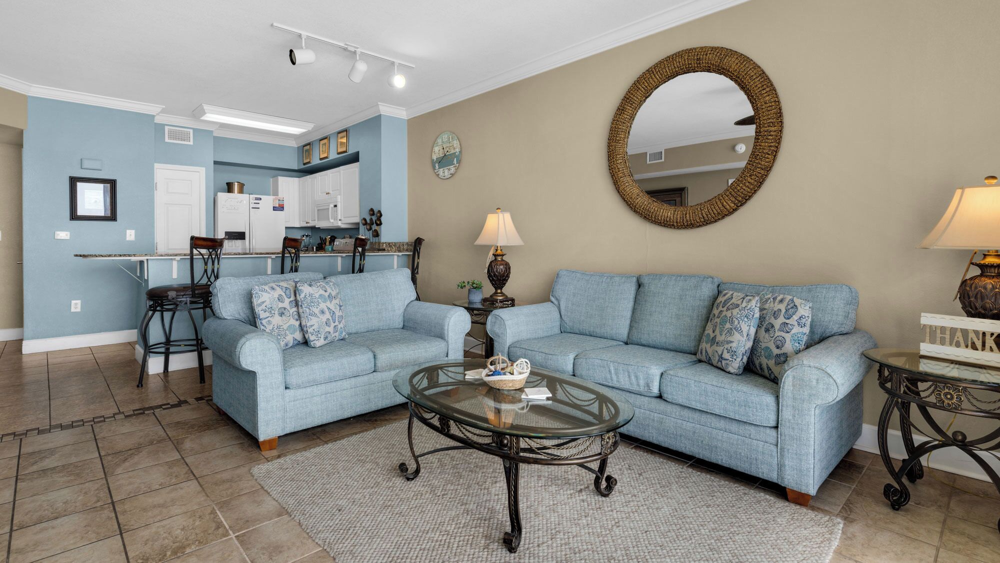 A living room with a blue sofa, a glass coffee table, and a kitchen in the background.
