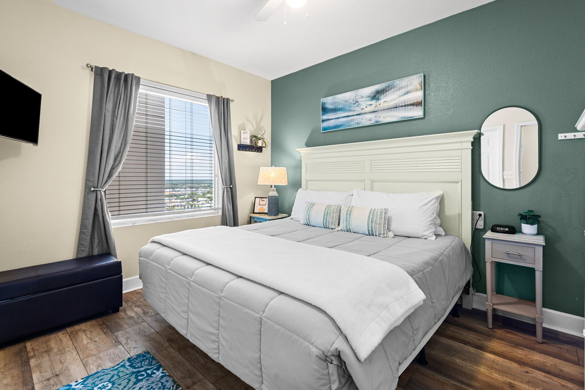 A bedroom with a bed, a bedside table, a lamp, a television, a window with curtains, and a wall-mounted mirror.