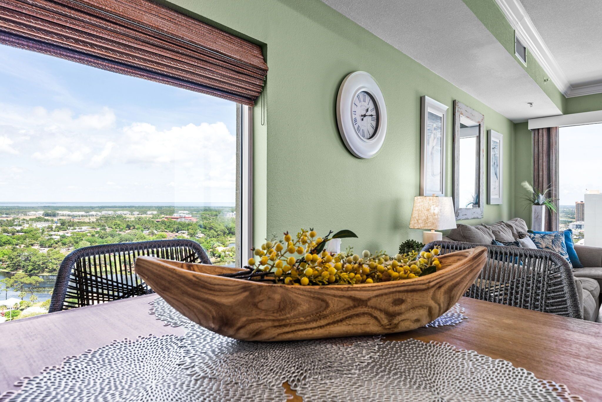 A dining area with a view of a landscape, a wooden bowl with yellow flowers, a clock, and a lamp.