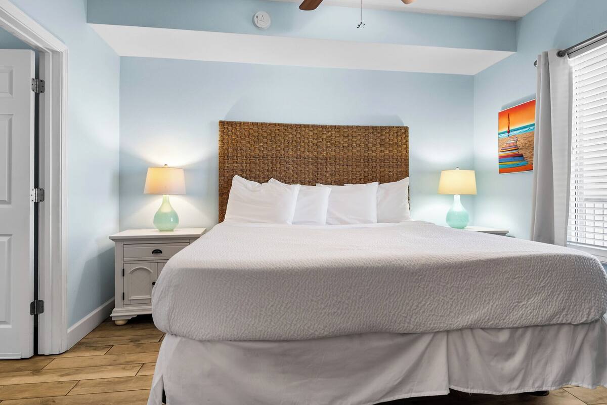 Una cama bien tendida con edredón blanco y almohadas, cabecera de fibra natural, dos lámparas de noche y un cuadro en la pared.