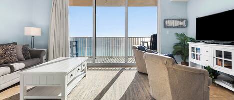 A living room with a sofa, a coffee table, a TV, and a balcony with a view of the sea.