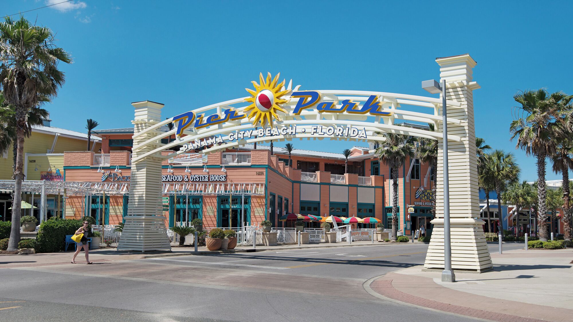 The entrance to Panama City Beach, Florida, featuring the iconic 'Panama City Beach' sign with a sun emblem, surrounded by palm trees and various beachfront restaurants.