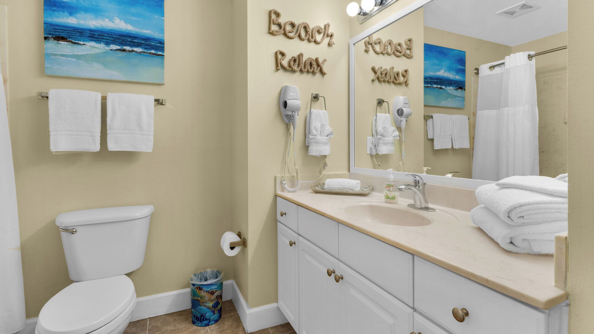 A bathroom with a toilet, sink, and mirror. There are towels hanging on the wall and a trash can.